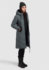 Grauer gesteppter Mantel mit Kapuze, Chevron-Steppung und Seitentaschen. Kombiniert mit einem schwarzen Oberteil, schwarzen Strumpfhosen und schwarzen Stiefeln.