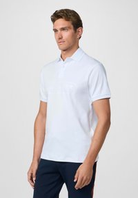 Biely polo tričko s jemne textúrovaným povrchom, klasickým golierom a krátkymi rukávmi. Na prednej strane je decentne potlačené logo.