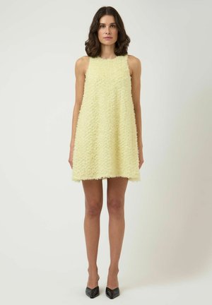 Kvinde stående med front mod kamera iført en ærmeløs, tekstureret, lysegul A-formet minikjole og sorte spidse hæle på en ensfarvet baggrund.