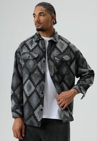 Man met gevlochten haar die een grijs-zwart jack met ruitpatroon draagt over een wit shirt, hij past het jack aan met zijn rechterhand.