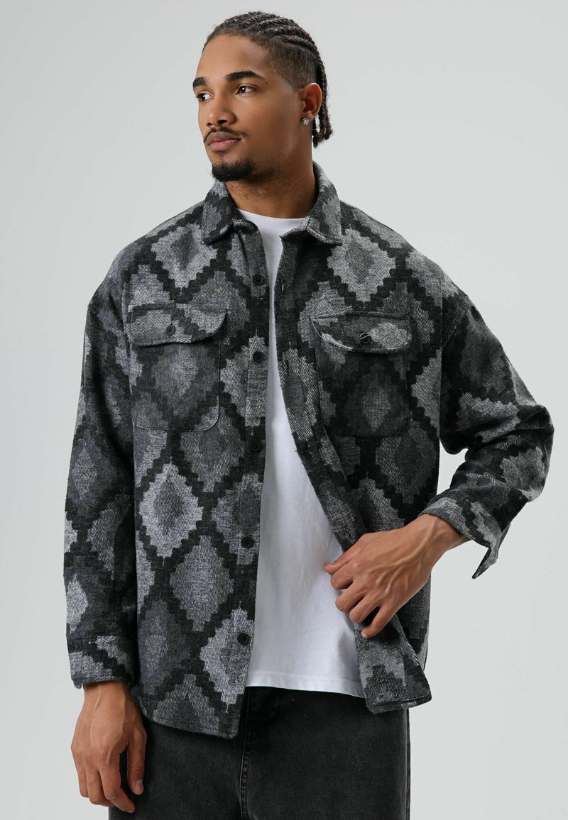 Man met gevlochten haar die een grijs-zwart jack met ruitpatroon draagt over een wit shirt, hij past het jack aan met zijn rechterhand.