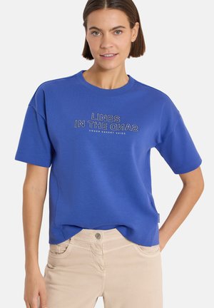Femme aux cheveux courts bruns portant un t-shirt bleu avec le texte "LINES IN THE SAND UNDER DESERT SKIES" et un pantalon beige, debout, une main sur la hanche.