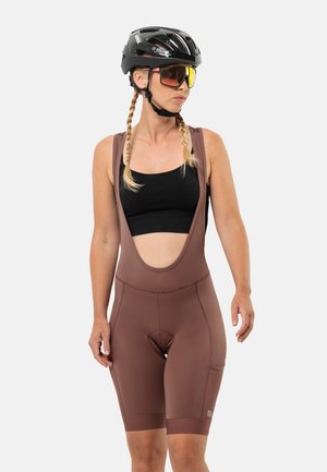 Brown cycling bib shorts with wide straps, a padded seat, and an open back design. Worn with a black sleeveless top and black helmet.