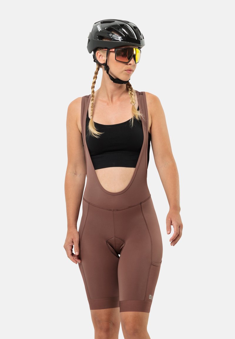 Bruine fiets-bibshorts met brede banden, een gevoerde zit en een open rugontwerp. Draag het met een zwart mouwloos shirt en een zwarte helm.