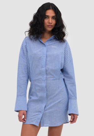 Kobieta o kręconych czarnych włosach, ubrana w niebiesko-białą, pionowo prążkowaną sukienkę-koszulę z szerokimi mankietami.