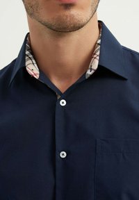 Chemise bleu marine à boutons avec un col à carreaux beige et noir, présentant une poche poitrine unique et des boutons blancs.