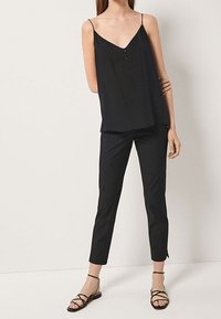 Vrouw in een zwart mouwloos V-hals top met dunne bandjes, slim fit zwarte cropped broek en zwarte platte sandalen met bandjes.