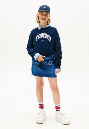 Sweatshirt marine avec le logo "TOMMY", chemise col en dessous de couleur bleu clair, jupe en denim bleu, baskets blanches et chaussettes rayées.
