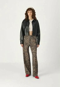 Mulher com cabelo encaracolado a usar uma camisa de couro preta cropped, uma t-shirt branca cropped, calças de padrão de leopardo, e sapatos vermelhos de bico fino, de pé sobre um fundo simples.