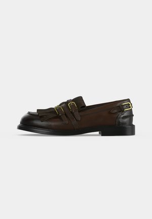 Mocasín de cuero marrón con puntera negra, doble hebilla dorada, detalle de borlas y tacón bajo negro sobre fondo blanco.