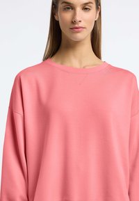 Jasnoróżowy sweatshirt z okrągłym dekoltem i subtelnymi przeszyciami. Luźny krój i miękka tekstura, z małym logo na ramieniu.