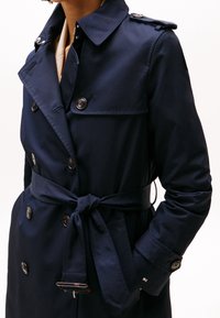 Tmavě modrý trenčkot z hladké látky, s dvouřadým zapínáním, pásem v pase a manžetami na knoflíky s detaily loga.