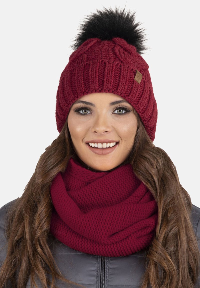 Gorro de punto burdeos con un pompón de piel sintética negra y una bufanda texturizada a juego, con un diseño acanalado y un aspecto acogedor.