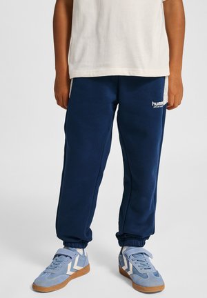 Granatowe spodnie dresowe z elastycznymi ściągaczami, z paskiem po boku i białym logo "hummel". Połączone z jasnoniebieskimi butami sportowymi.