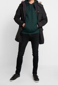 Parka negra con capucha forrada de pelo, sudadera verde y jeans negros. Zapatillas negras con tres correas. La parka cuenta con bolsillos.