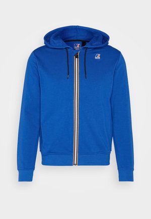 Sudadera con capucha azul real con cremallera, cordones negros, cremallera a rayas multicolor y pequeño logo en el pecho y etiqueta interior del cuello.