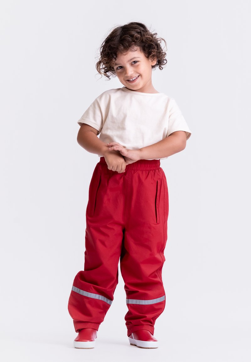 Rote wasserdichte Hosen mit reflektierenden Streifen und Seitentaschen, kombiniert mit einem cremefarbenen Kurzarmhemd und roten Schuhen.