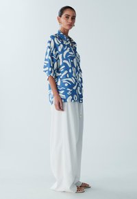 Blue and white patterned blouse with a relaxed fit, paired with lightweight, flowing white trousers and flat sandals.