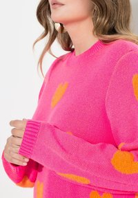 Różowy sweter z miękkiego materiału, zdobiony dużymi pomarańczowymi sercami, z prążkowanymi mankietami i okrągłym dekoltem. Szczegóły ukazują fakturę i kontrast kolorów.