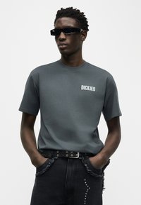 Άνδρας που φοράει σκούρο γκρι μπλουζάκι Dickies, μαύρη ζώνη με τρουκς, μαύρα γυαλιά ηλίου και μαύρο τζιν με φθαρμένες τσέπες και διακοσμητικά τρουκς.