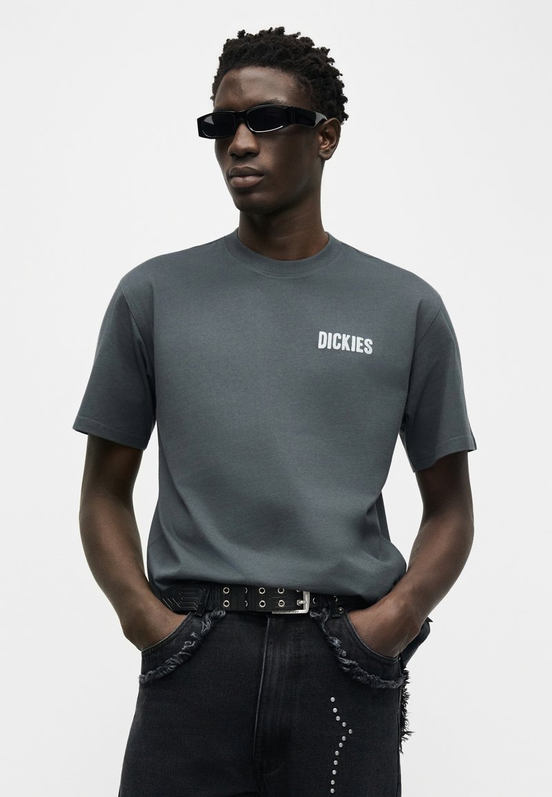 Hombre con camiseta Dickies gris oscuro, cinturón negro con tachuelas, gafas de sol negras y vaqueros negros con bolsillos deshilachados y tachuelas decorativas.