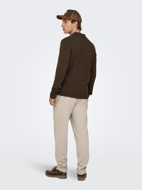 Pullover marrone e pantaloni della tuta beige abbinati a scarpe marroni, con un cappello marrone e beige. I materiali sembrano morbidi e casual.