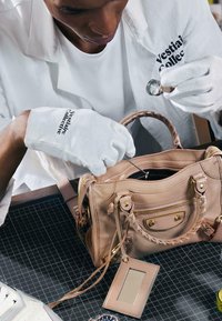 Personne portant des gants blancs examine un sac à main en cuir beige avec une loupe et un outil sur un tapis en grillage noir.