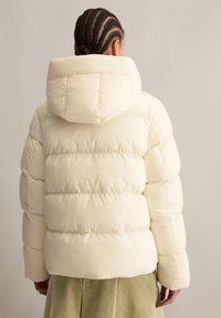 Cremfarbener Pufferjacke mit Kapuze, die über gesteppten Abschnitten und einer glatten, glänzenden Textur verfügt. Das Design ist kurz und hat eine taillierte Silhouette.