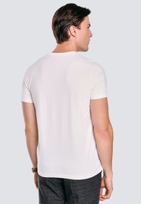 Baltas trumpas rankovėmis marškinėliai su apvalia iškirpte, minkšta tekstūra ir paprastu dizainu, matomi iš nugaros prieš neutralų foną.