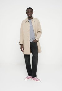 Μπεζ παλτό τύπου trench με καφέ κουμπιά, φορεμένο πάνω από γκρι πουκάμισο, σε συνδυασμό με μαύρο παντελόνι και ροζ αθλητικά παπούτσια. Απλός σχεδιασμός, άνετη εφαρμογή.