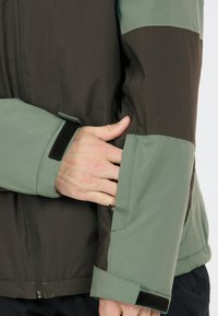 Grüne und braune Jacke mit Reißverschluss, verstellbaren Klettverschluss-Manschetten und auffälligem Farbabstufungsdesign. Glatte Textur, taillierte Passform.