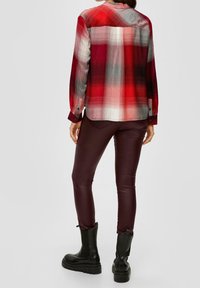 Camisa de cuadros roja y negra con mangas largas, detalles de botones, llevada con pantalones burdeos y botas negras. Tejido suave y corte relajado.