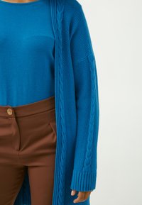 Lungo cardigan blu a maglia con motivo a treccia lungo i bordi, abbinato a una maglietta blu e a un pantalone marrone a vita alta con dettaglio di bottone dorato.