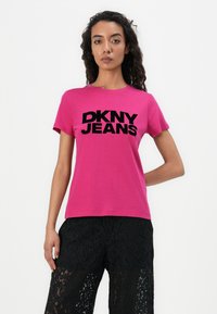 Vaaleanpunainen puuvillapaita, jossa on musta "DKNY JEANS" -logo. Lyhyet hihat, pyöreä pääntie, yhdistetty mustiin pitsiraidoitettuihin housuihin.