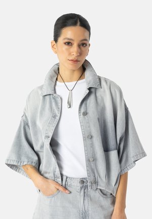Giovane donna con capelli raccolti che indossa una giacca di jeans oversize grigio chiaro, pantaloni abbinati, maglietta bianca e una collana con ciondolo d'argento.