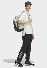 Mochila con tela gris y detalles en negro, usada sobre una camiseta blanca, combinada con pantalones negros con tres franjas blancas y zapatos negros.