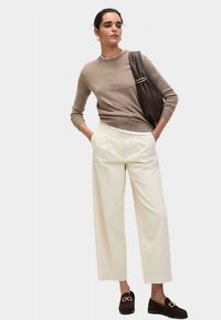 Brązowy sweter z dzianiny, białe spodnie z wysokim stanem i ciemne mokasyny. Duża brązowa torba ze złotym akcentem. Prosty nowoczesny design.