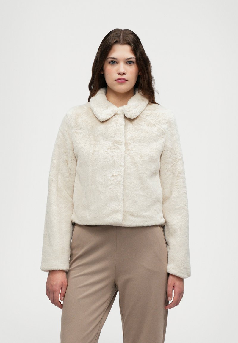 Krem jaka od lažnog krzna s okruglim ovratnikom, dugim rukavima i skraćenim dizajnom. Glatka tekstura bez vidljivih zatvarača ili uzoraka.