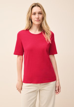 T-shirt rossa a maniche corte con scollatura rotonda, realizzata in un tessuto morbido. Il modello la indossa con pantaloni chiari su uno sfondo neutro.