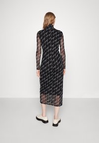 Femme aux longs cheveux portant une robe midi noire en tissu transparent imprimée du texte blanc "Hugo" en répétition, associée à des chaussures couleur crème.