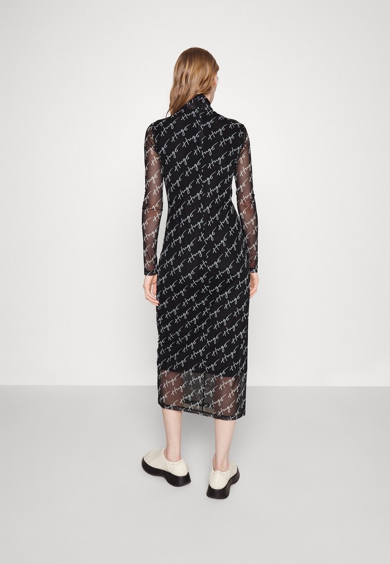 Femme aux longs cheveux portant une robe midi noire en tissu transparent imprimée du texte blanc "Hugo" en répétition, associée à des chaussures couleur crème.