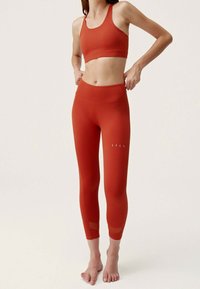 Červený sportovní crop top a legíny s vysokým pasem, oba s hladkým povrchem. Legíny mají průhledné síťované detaily na lýtkách.