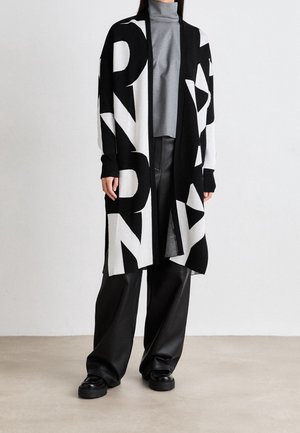 Personne portant un long cardigan noir et blanc ouvert avec des lettres en gras, un col roulé gris, un pantalon large noir et des chaussures noires sur un fond neutre.