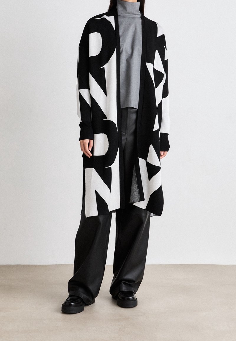 Personne portant un long cardigan noir et blanc ouvert avec des lettres en gras, un col roulé gris, un pantalon large noir et des chaussures noires sur un fond neutre.
