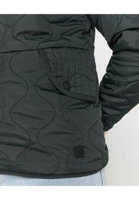 Veste matelassée noire avec un motif de vague texturé, une grande poche à rabat avec une fermeture à pression et une étiquette.