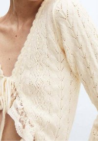 Jasnokremowy, dziergany sweter na guziki z wzorem diamentowym, falistymi krawędziami i koronkową lamówką u dołu. Posiada zapięcie na tęczkę z przodu.