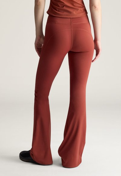 Bagfra billede af person iført rustfarvede viddebukser og matchende top med sorte sko, stående på neutral baggrund.