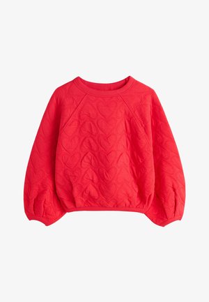 Jeune fille aux cheveux bouclés porte un pull matelassé rouge, des leggings blancs avec des cœurs rouges et des baskets montantes noires, debout sur fond beige.