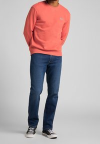 Koraal sweatshirt, ronde hals, ribgebreide manchetten en zoom; gedragen met blauwe jeans en zwarte sneakers. Opvallend logo op de borst.