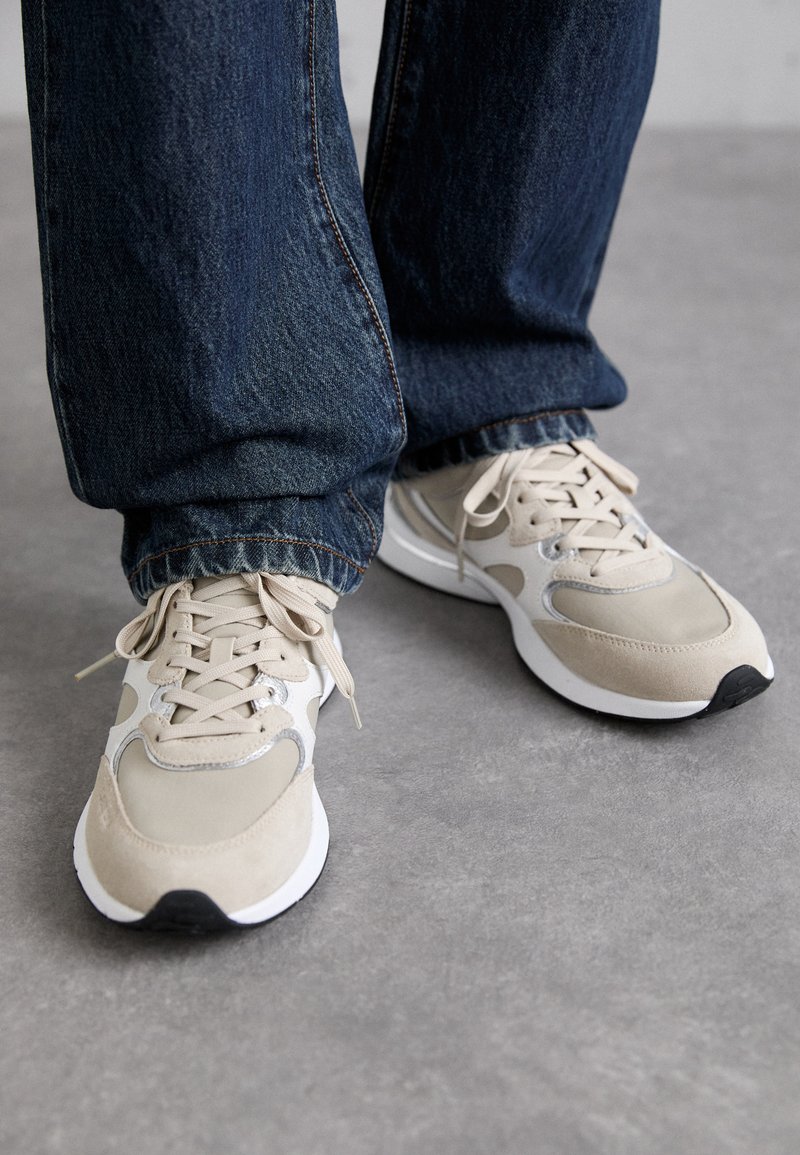 Sneakers beige e grigi con un mix di materiali in suede e mesh, dotati di chiusura con lacci e una suola bianca con un accento nero nella punta.